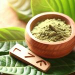 Kratom powder in wooden bowl surrounded by kratom leaves.